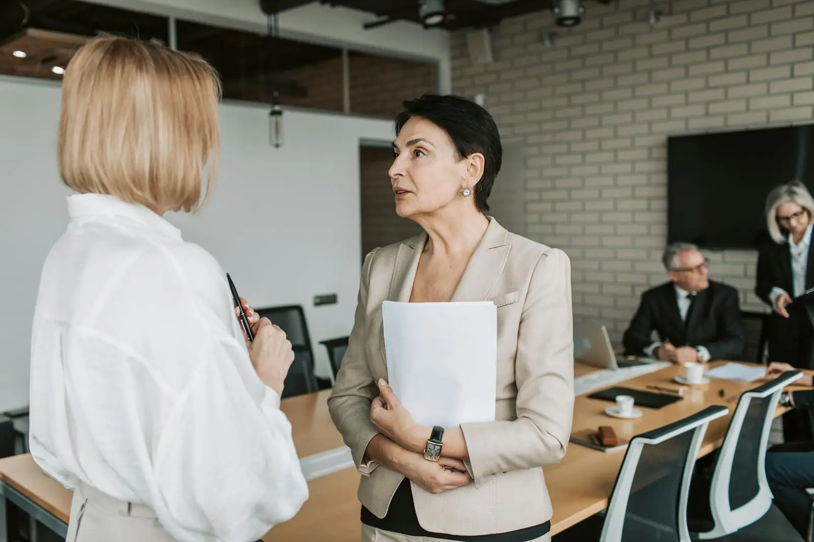 Two women in conversation in a warm office, with a team meeting behind them